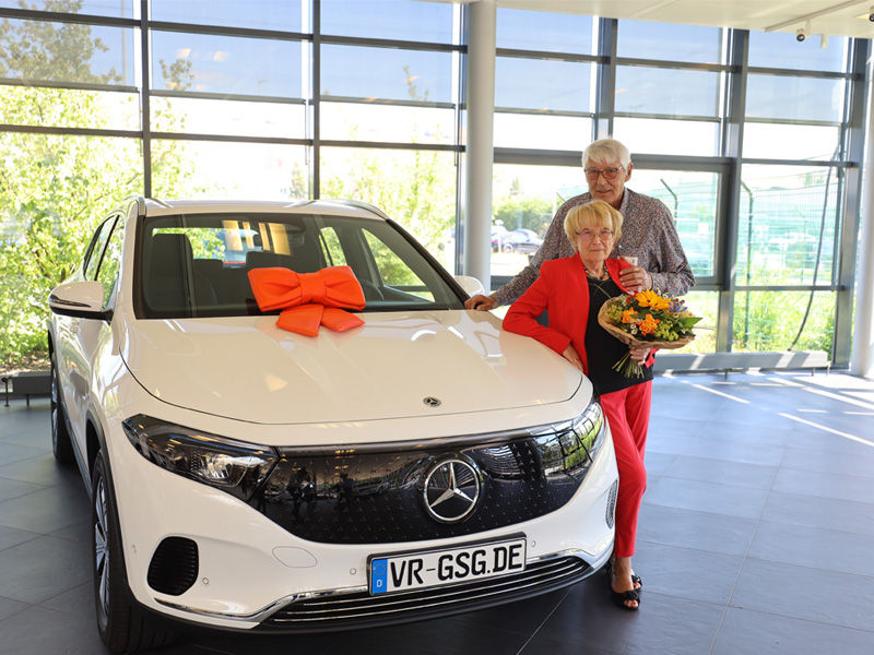 Ein älteres Ehepaar steht mit einem Blumenstrauß in der Hand rechts neben einem Mercedes-Benz EQA.