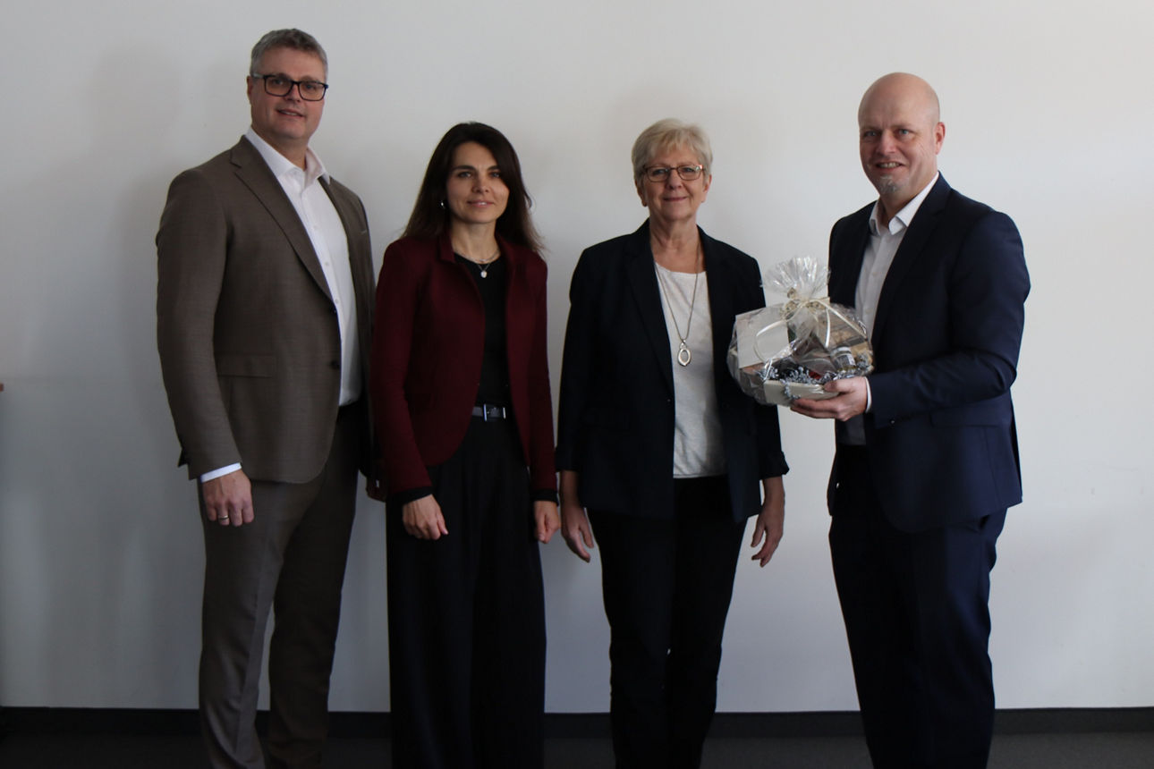 Auf dem Bild stehen vor einer weißen Wand zwei Männer und zwei Frauen. Einer der Männer hält einen Präsentkorb in der Hand. 