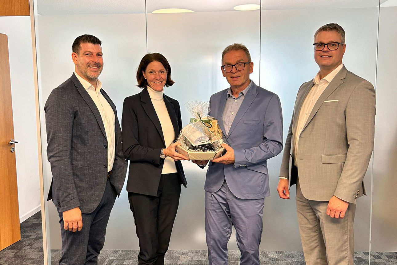 Drei Männer und eine Frau stehen in einem hellen Raum. Die Frau überreicht einen Präsentkorb an einen der Männer, den Jubilar.. 