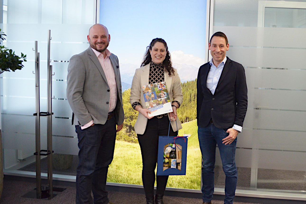 Mitarbeiterin steht mit Geschenkkorb neben beiden Vorständen in der Filiale vor einer Glasstrennwand