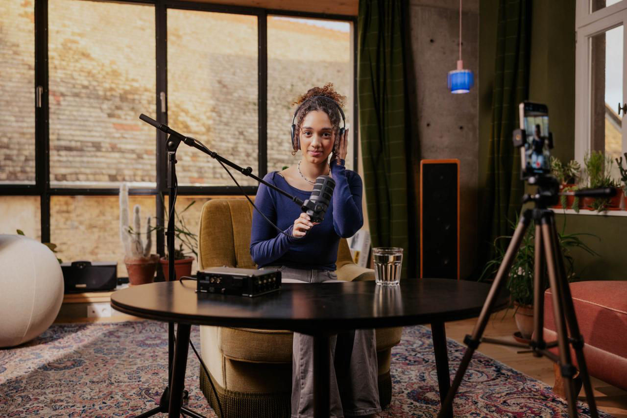 Junge Frau mit Kopfhörern spricht in einem gemütlichen Studio vor Mikrofon und Kamera.