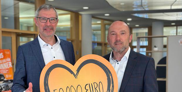 Zwei Männer in Anzügen zeigen stolz ein Schild mit der Aufschrift „500.000 Euro“.