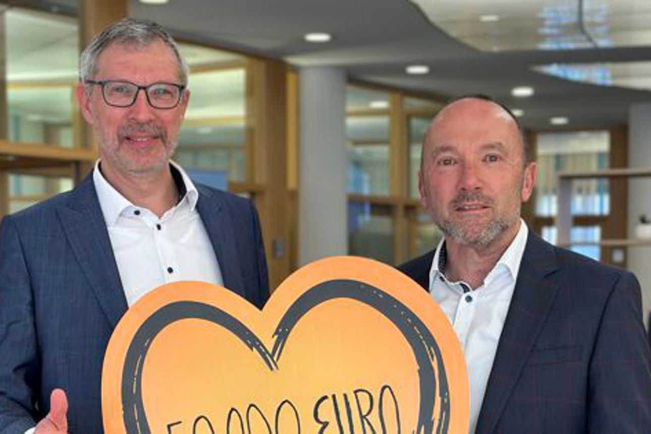 Zwei Männer in Anzügen zeigen stolz ein Schild mit der Aufschrift „500.000 Euro“.