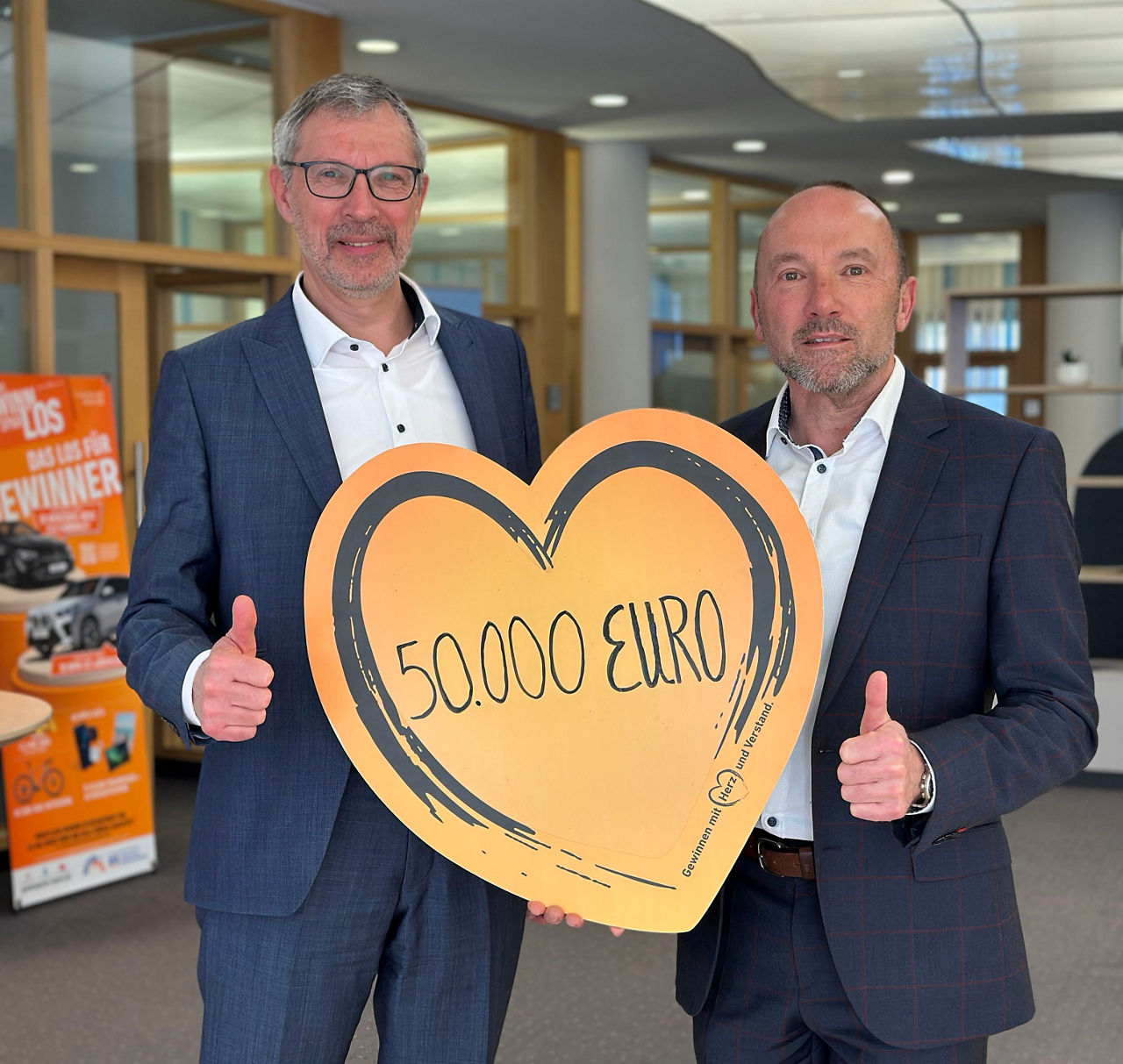 Zwei Männer in Anzügen zeigen stolz ein Schild mit der Aufschrift „500.000 Euro“.