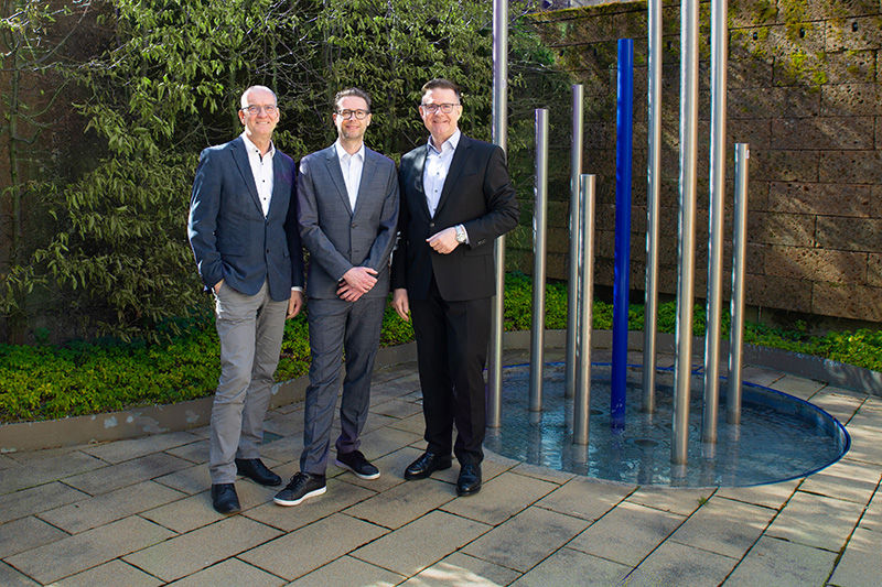 Vorstandssprecher Herr Schöpfel und die Mitarbeiter Herr Rohrmüller und Herr Richter vor einer Brunnenskulptur