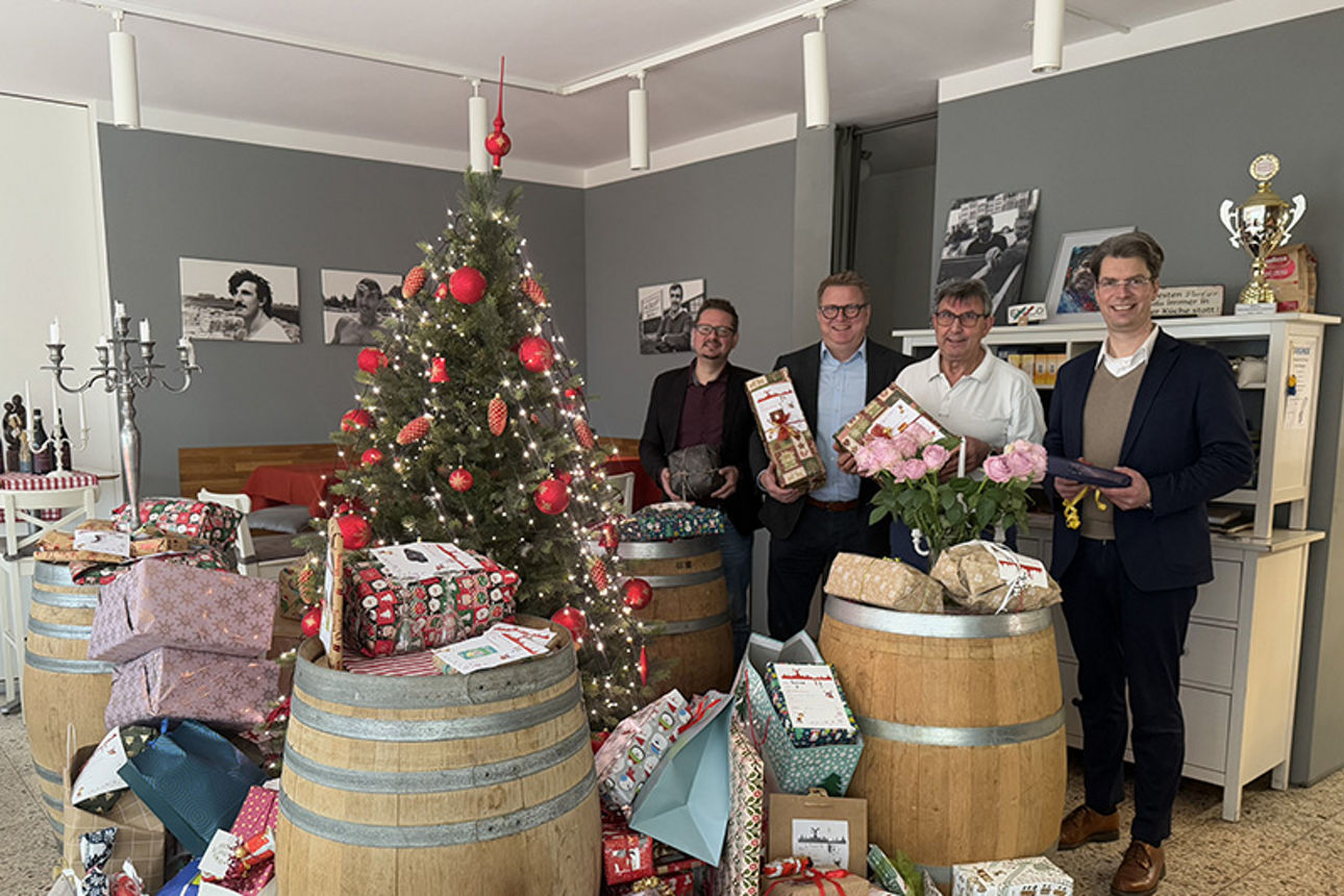 Vier Männer stehen neben einem Weihnachtsbaum, vor dem viele Geschenke liegen, und halten auch Geschenke in der Hand.
