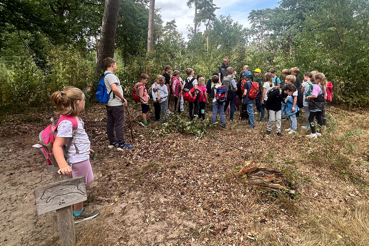 Eine Gruppe Kinder steht vor einer Frau und lauscht Ihr.