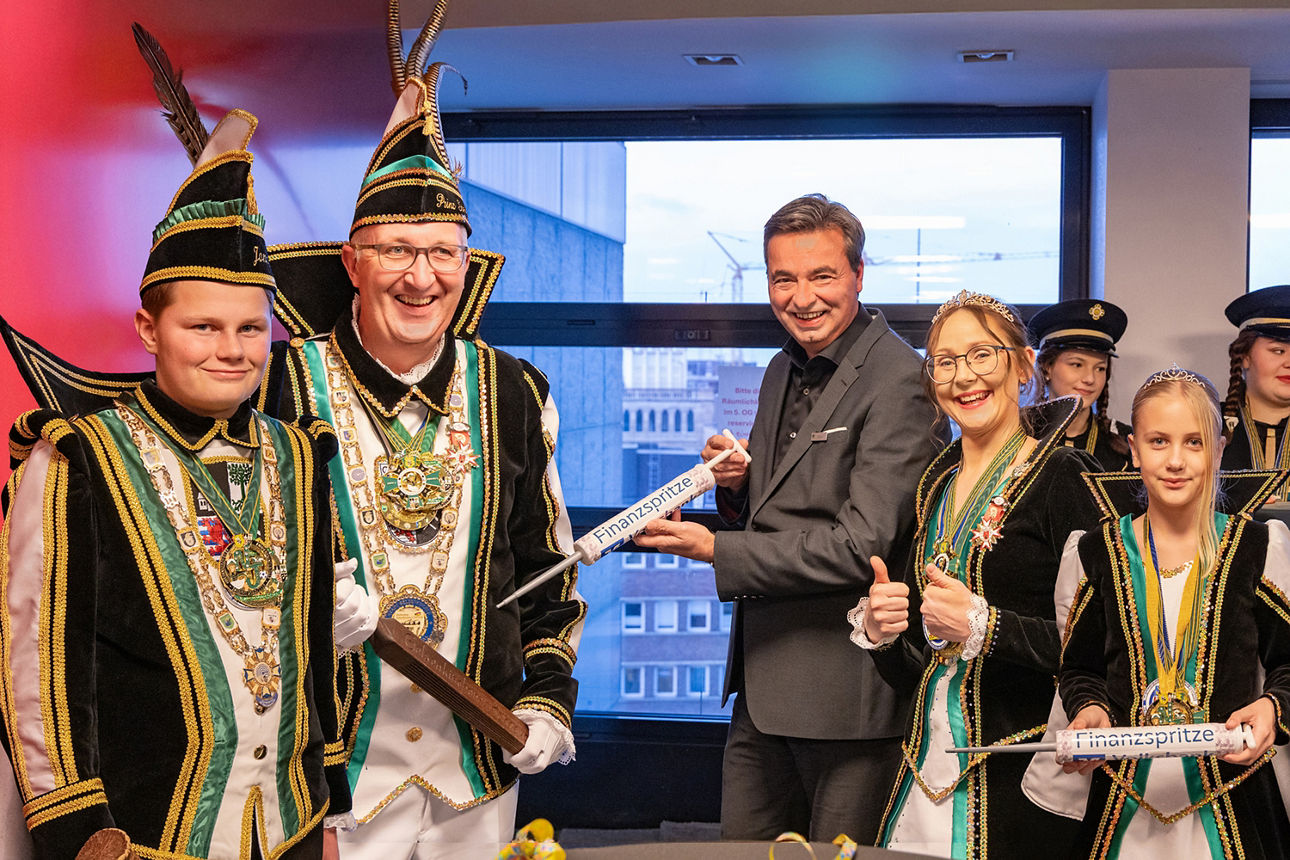 Zu sehen sind sowohl das Stadtprinzenpaar des KG Piccolo als auch das Kinderprinzenpaar aus dem selben Karnevalsverein, alle im Ornat. Dazwischen steht lächelnd unser Vorstand Jörg Lott und hält symbolisch eine übergroße Spritze mit der Aufschrift "Finanzspritze" in Rirchtung Stadtprinz.