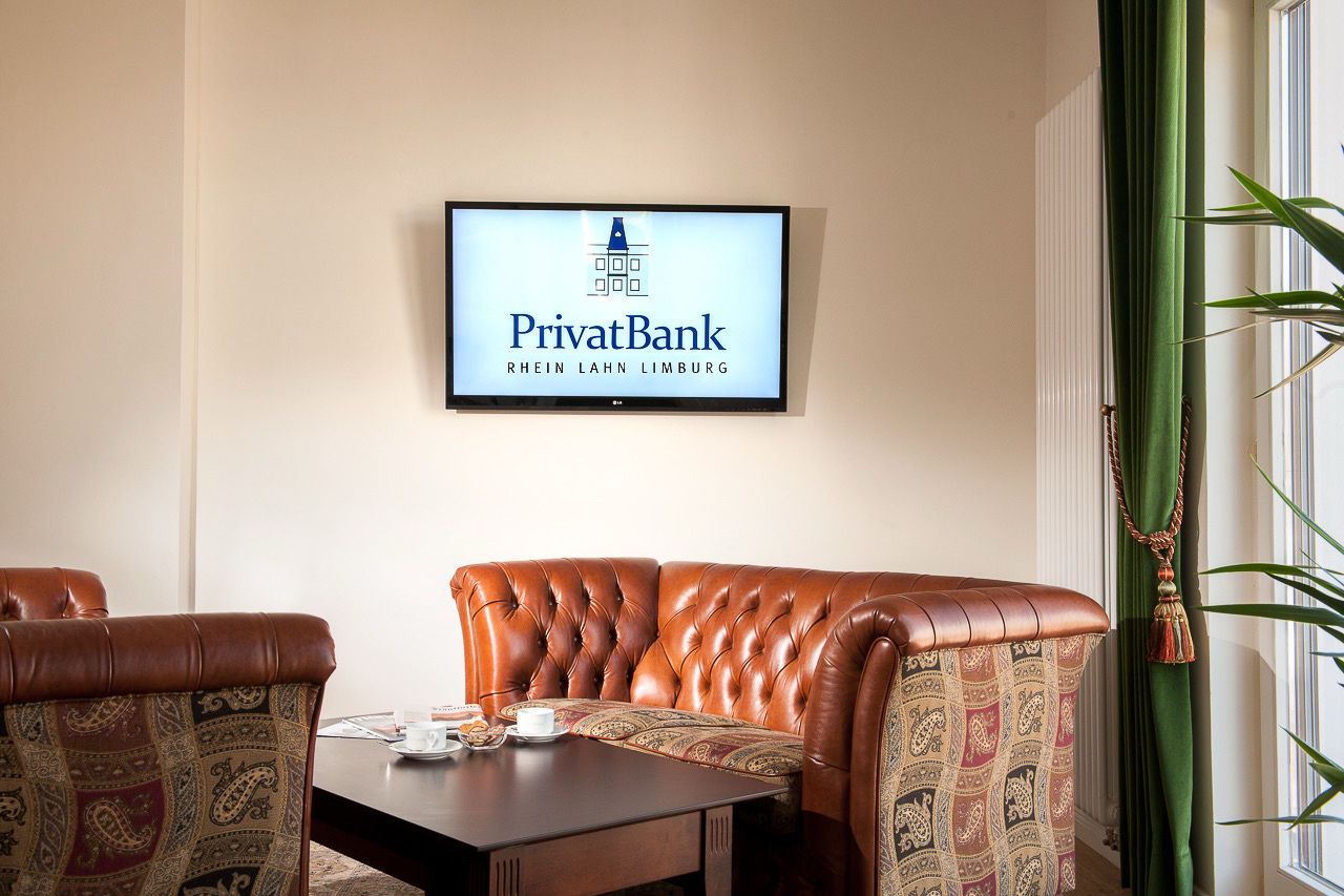 Ein gemütlicher Sessel, der vor einer beigen Wand steht, an der ein Bildschirm mit dem Logo der PrivatBank Rhein Lahn Limburg hängt.