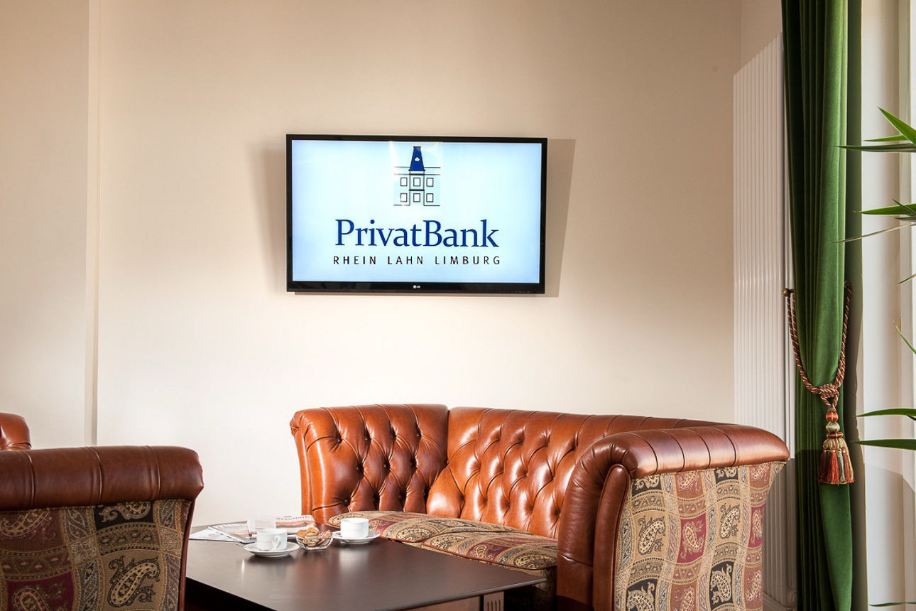 Ein gemütlicher Sessel, der vor einer beigen Wand steht, an der ein Bildschirm mit dem Logo der PrivatBank Rhein Lahn Limburg hängt.
