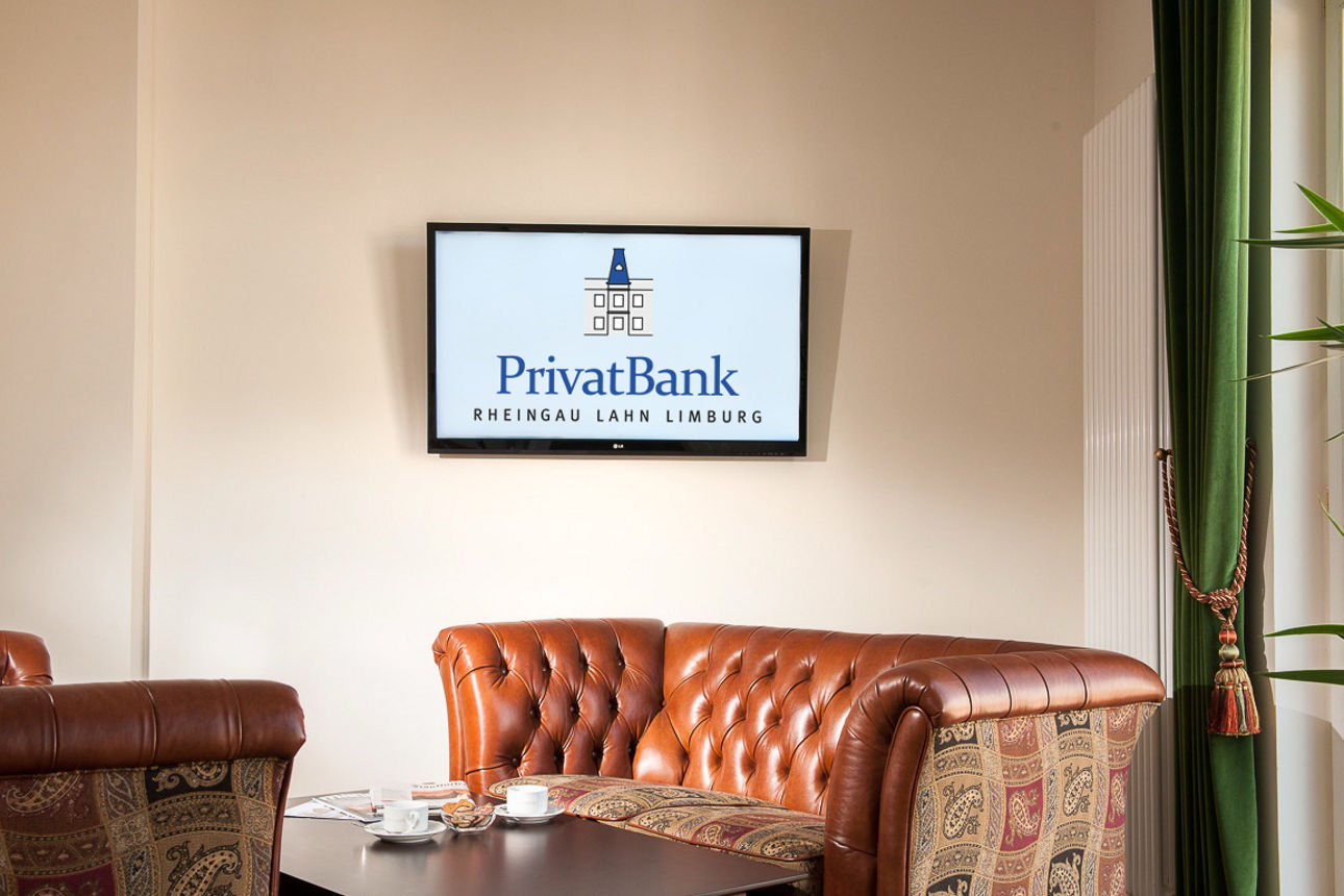 Ein gemütlicher Sessel, der vor einer beigen Wand steht, an der ein Bildschirm mit dem Logo der PrivatBank Rhein Lahn Limburg hängt.