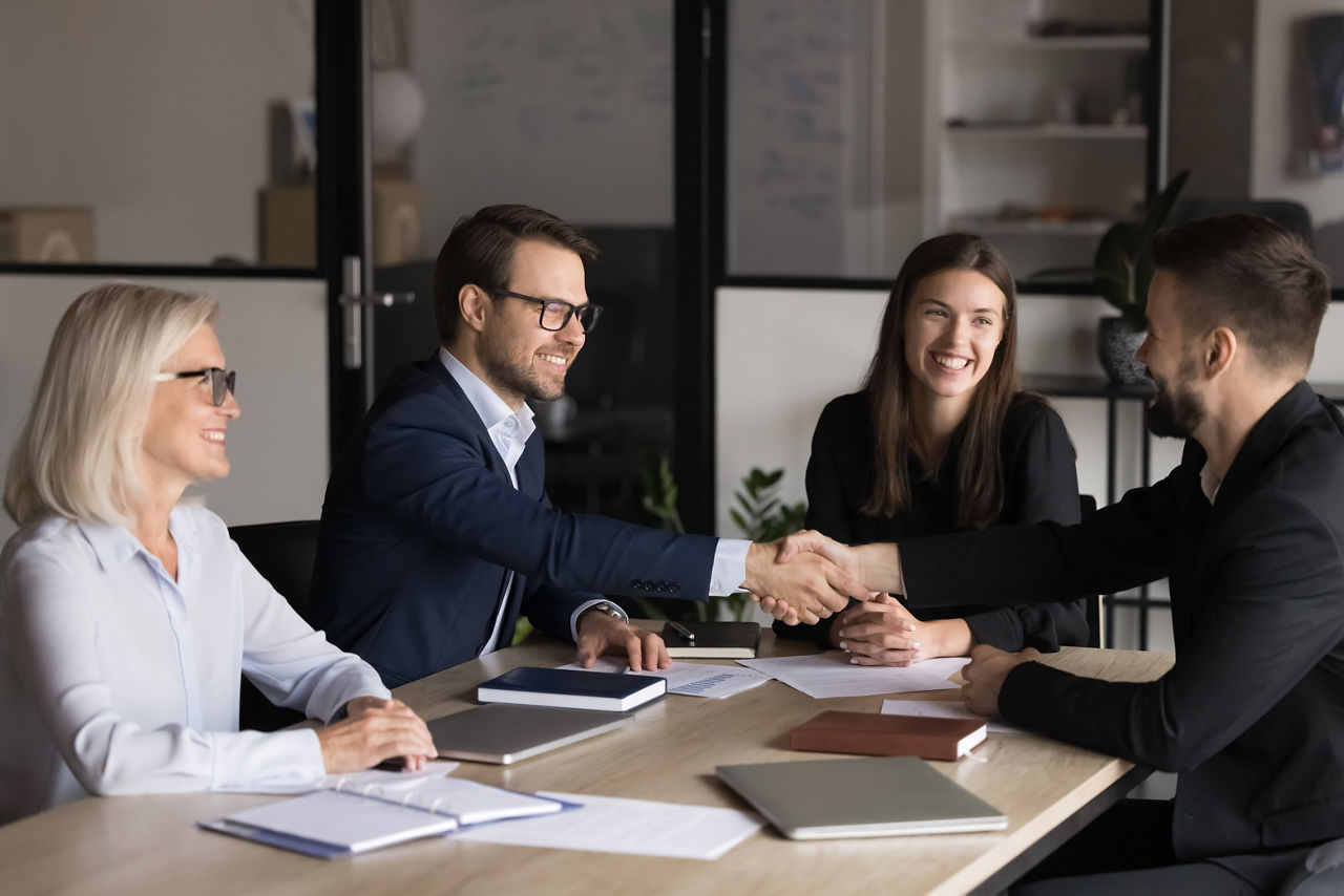 Zwei Männer und zwei Frauen sitzen in einem Büro an einem Tisch. Die beiden Männer geben sich die Hand.