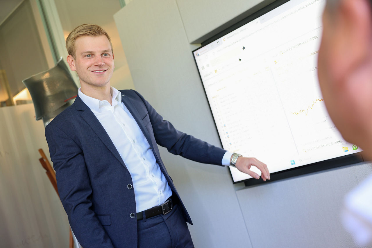 Lucas Elsbecker steht an einem großen TV-Bildschirm und präsentiert einen Graphen der Börse