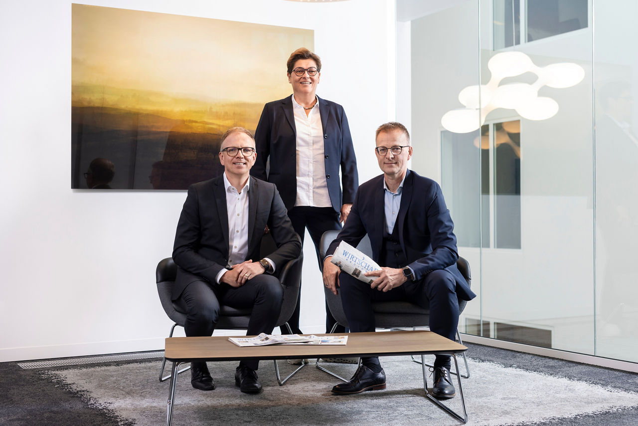 Eine Frau und zwei Männer die direkt in die Kamera blicken. Im Hintergrund ein großer Kunstdruck. 