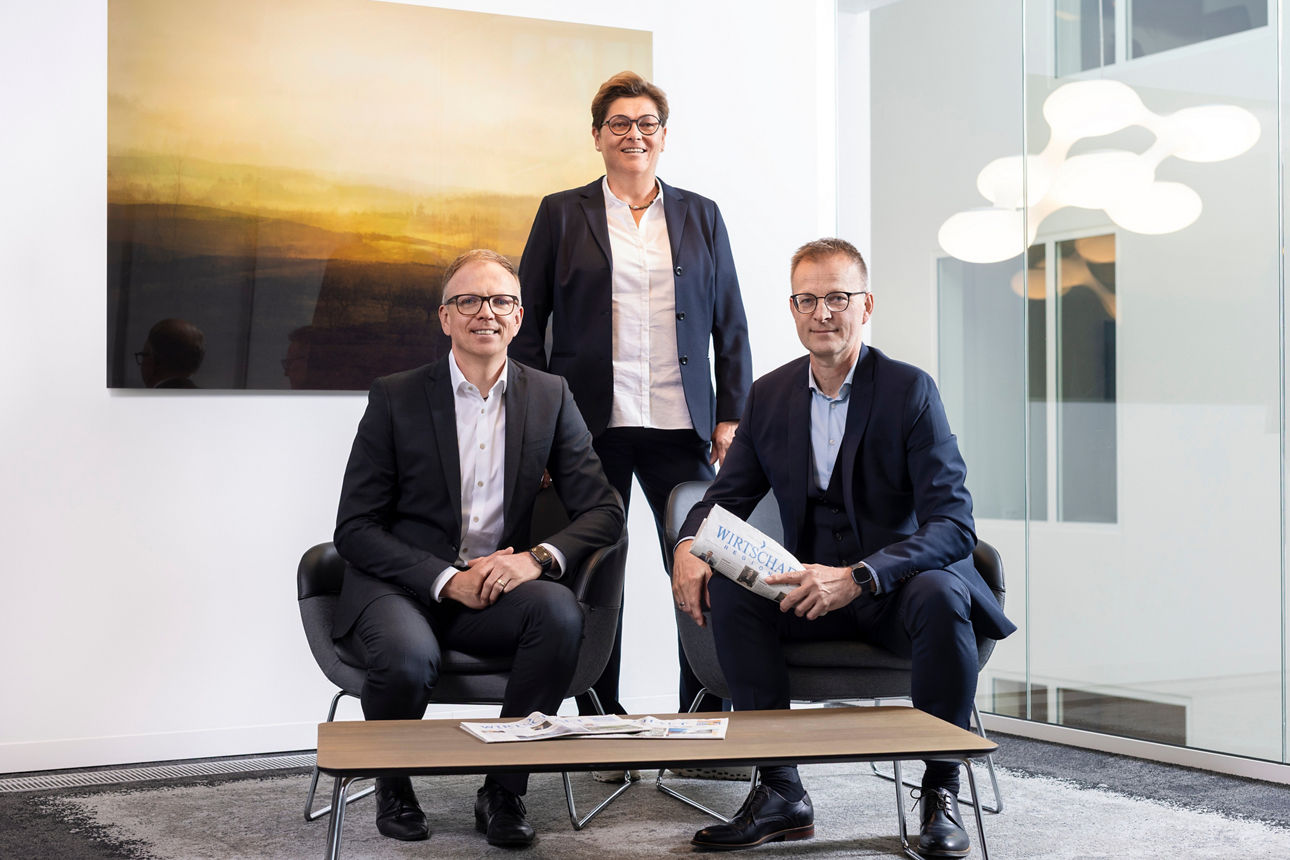 Eine Frau und zwei Männer die direkt in die Kamera blicken. Im Hintergrund ein großer Kunstdruck. 