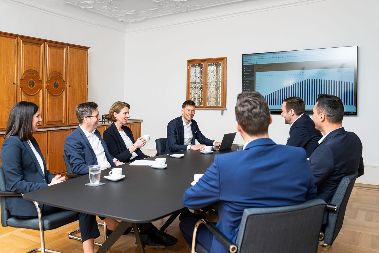 Die Private Banking Berater sitzen um einen Tisch bei einer Besprechung - im Hintergrund ist ein Fernseher mit einer Präsentation