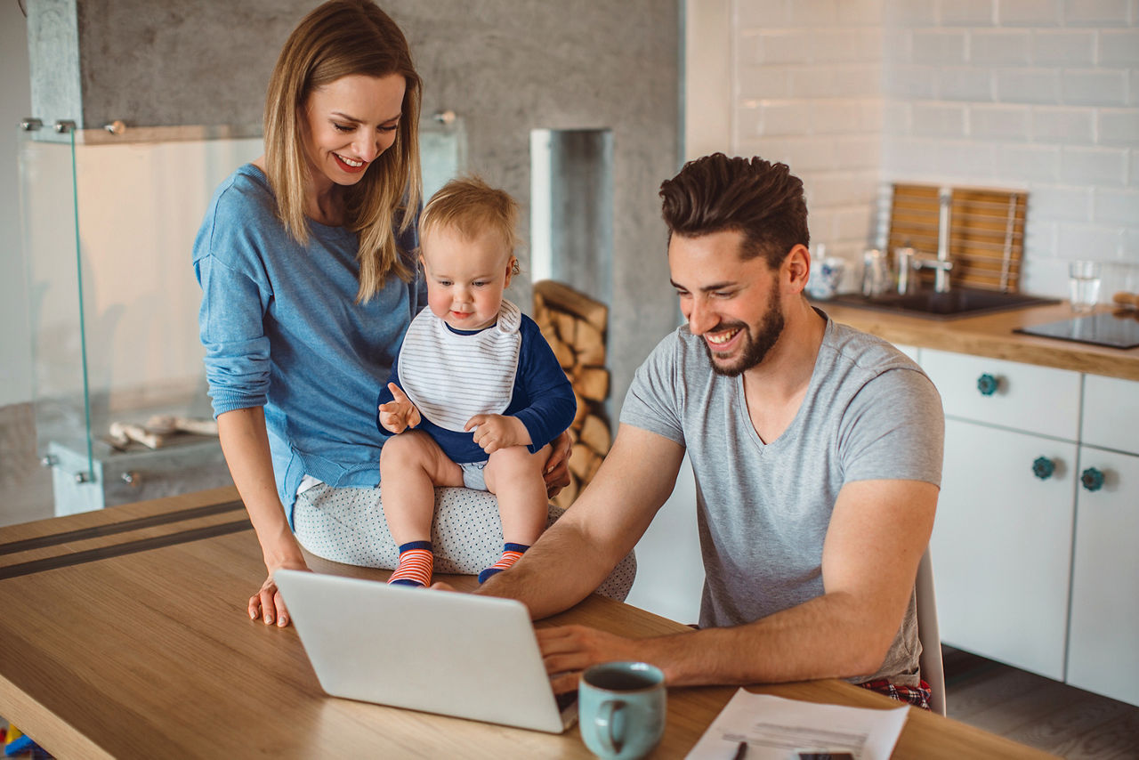 Am Küchentisch sitzt ein junger Vater am Laptop. Neben ihm sitzt seine blonde, junge Frau halb auf der Tischplatte und hat dabei das gemeinsame Baby auf dem Schoß. Alle lächeln fröhlich und schauen auf das Display des Laptops.