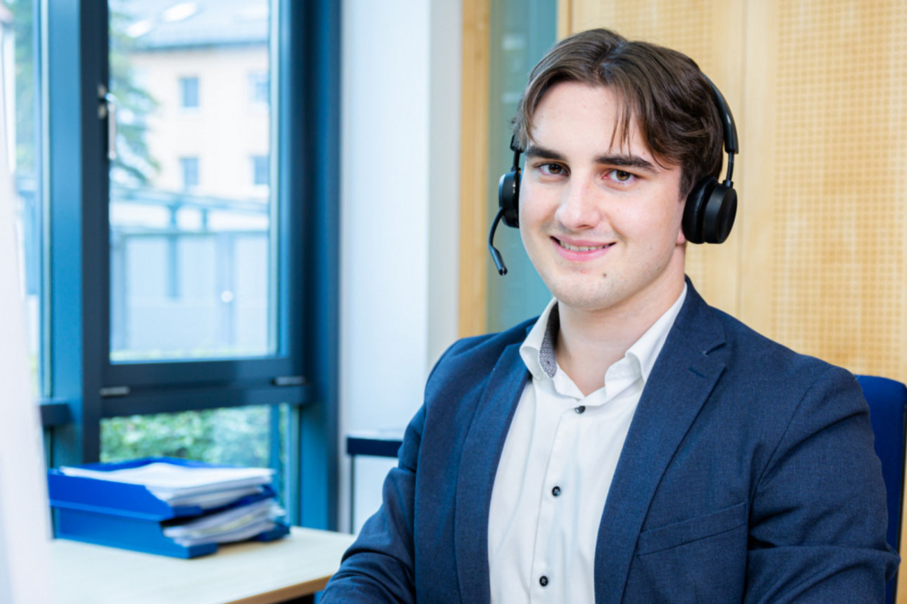 Mann mit Headset sitzt lächelnd im Büro vor Regalen und Computerarbeitsplatz.