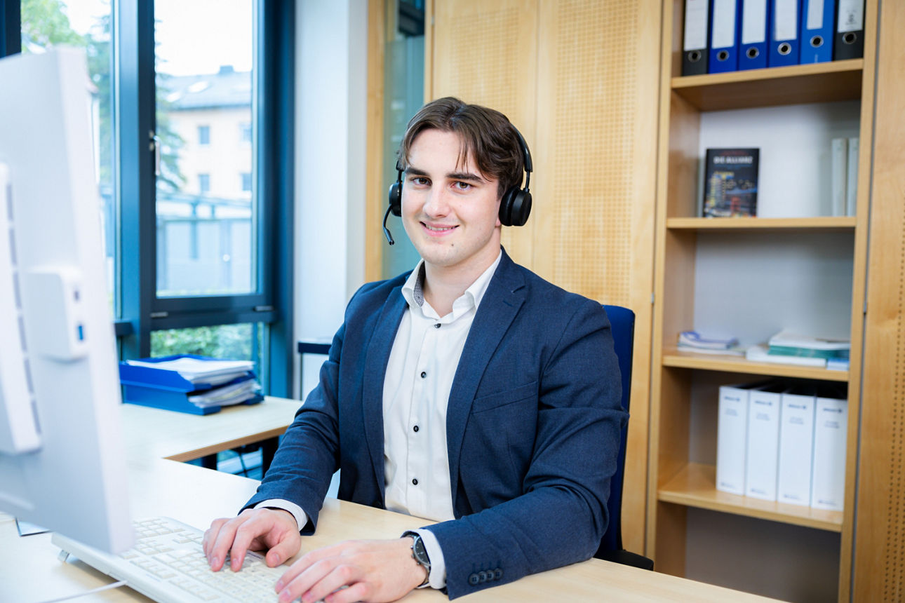 Mann mit Headset sitzt lächelnd im Büro vor Regalen und Computerarbeitsplatz.