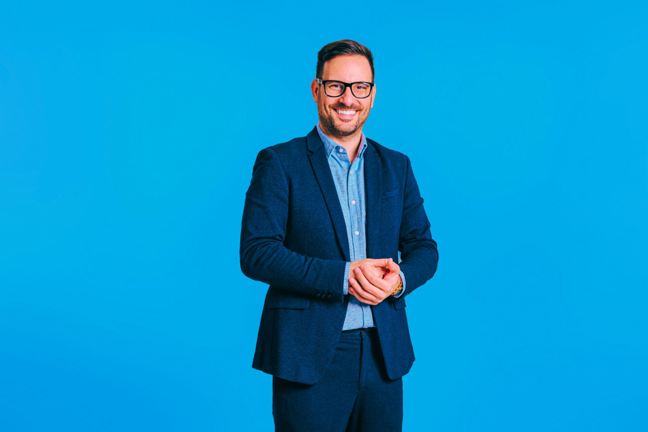 Lächelnder Mann mit Brille in blauem Anzug steht mit gefalteten Händen vor blauem Hintergrund