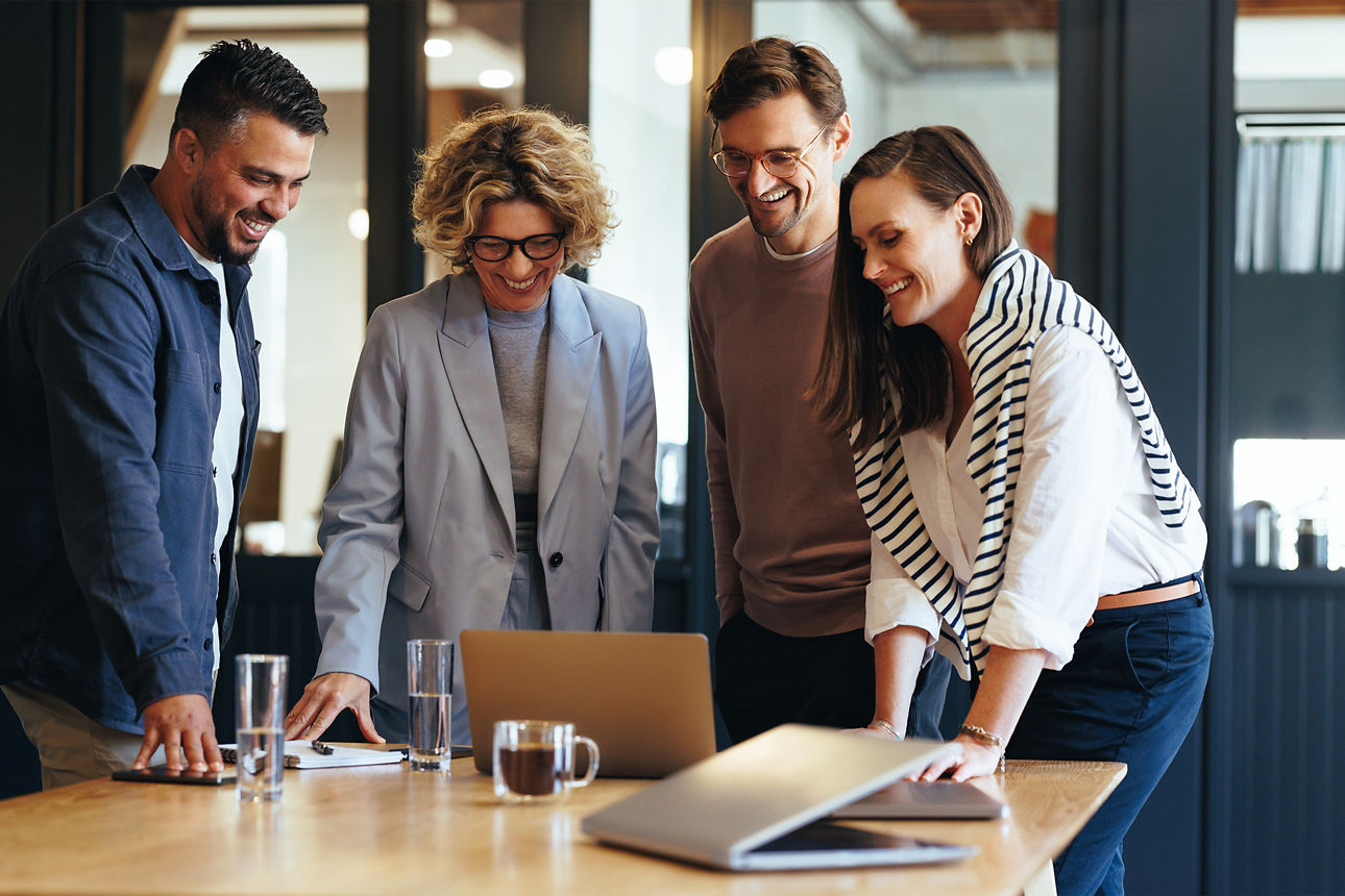 Besprechungssituation. Zwei Männer und zwei Frauen stehen im Halbkreis und schauen lächeln auf einen Laptop.