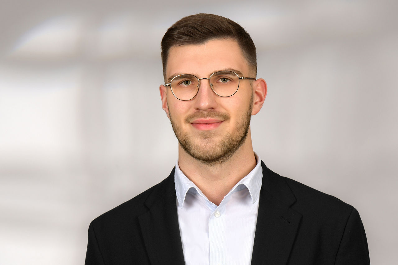 Mann mit kurzem Haar und Brille trägt schwarzen Anzug und weißes Hemd vor unscharfem Hintergrund