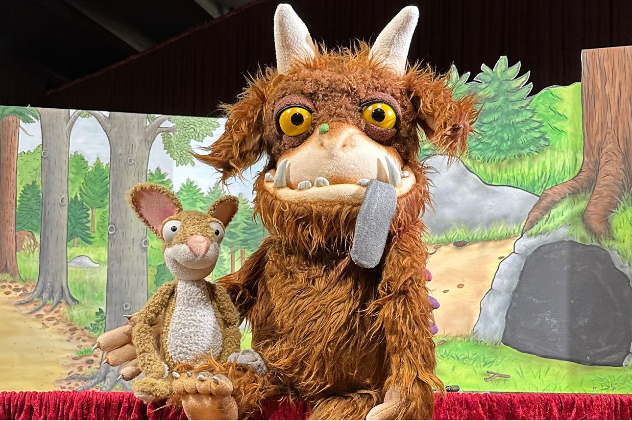 Braunes, zotteliges Monster mit großen gelben Augen und zwei weißen Hörnern neben einem braunen, sitzenden Hasen mit großen Ohren vor Waldkulisse.