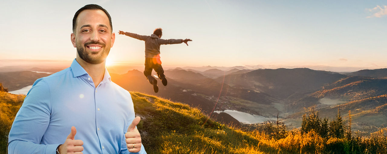 Ein junger Mann mit zwei hochgestreckten Daumen. Im Hintergrund macht ein Mann einen Luftsprung in den Bergen