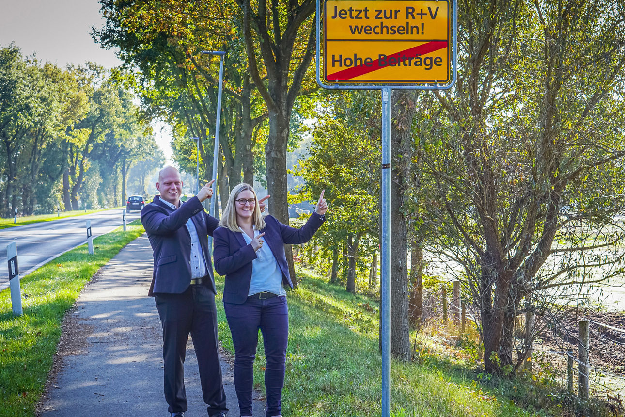 Zwei Personen stehen lächelnd an einer Landstraße und zeigen auf ein gelbes Ortsschild mit Werbung und dem durchgestrichenen 