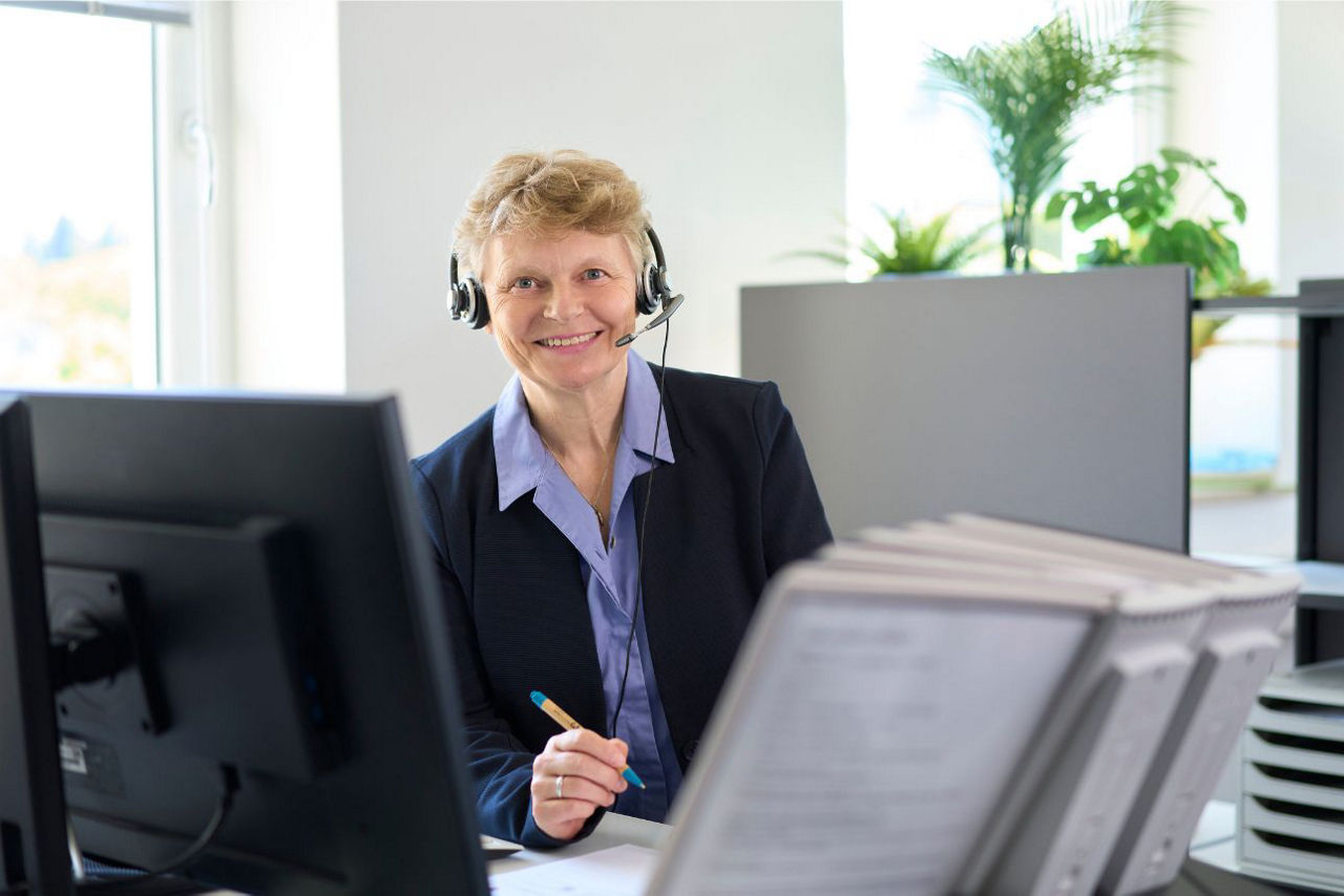 Lächelnde Frau in Business-Bekleidung steht an mit Headset an ihrem Büroarbeitsplatz