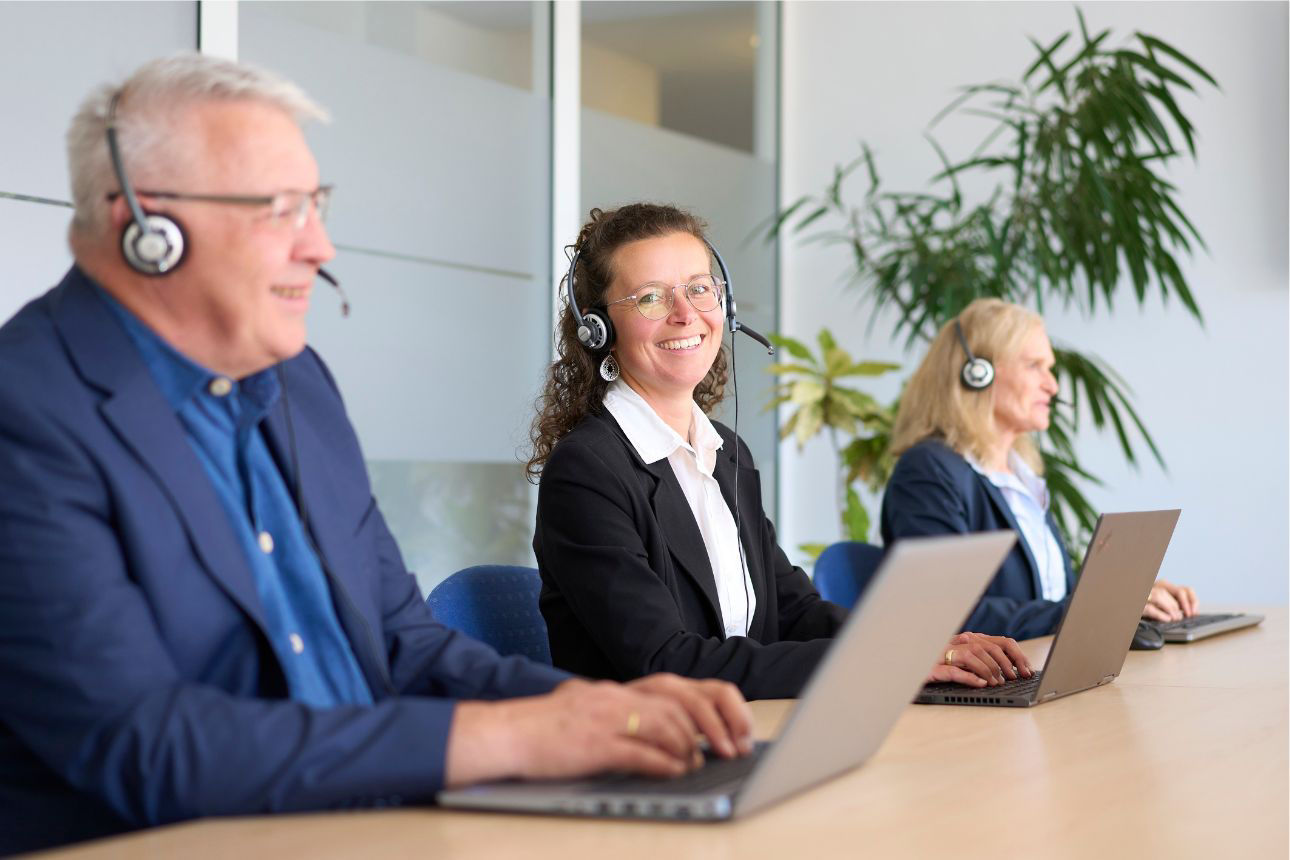 Drei lächelnde Personen (zwei Frauen, ein Mann) mit Headsets, die an einem langen Tisch sitzen und an Laptops arbeiten.