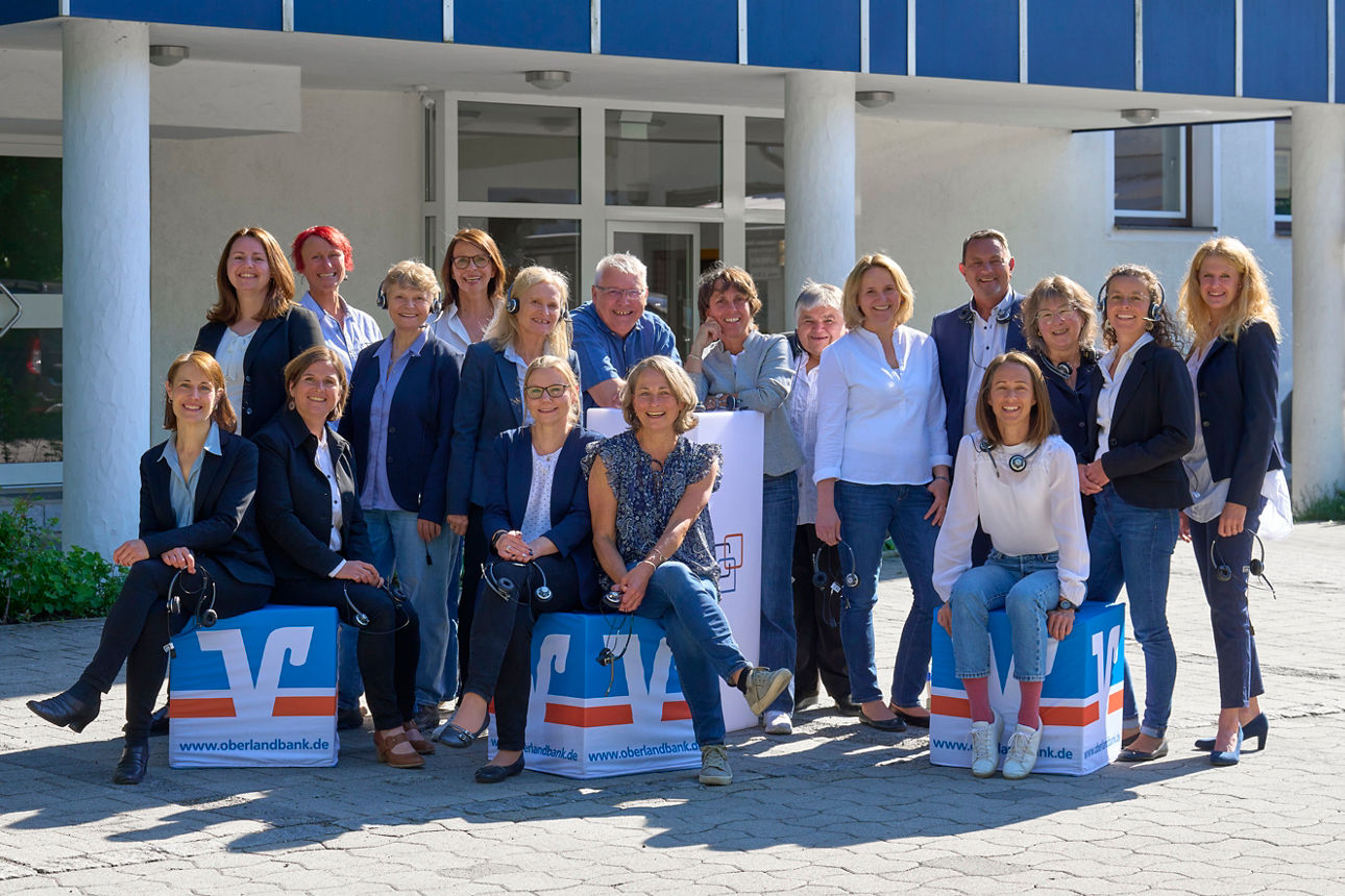 Eine große Gruppe von Mitarbeitern des KST (Kundenservice-Teams) der RBimOberland, einige mit Headsets, posieren draußen vor einem Gebäude, einige sitzen auf blauen Kisten mit dem Logo der Bank.