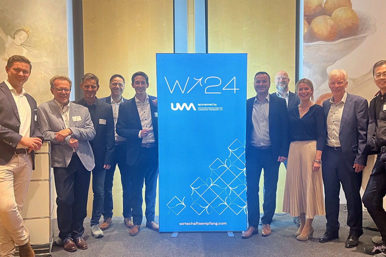 Eine Gruppe von neun Personen, hauptsächlich Männer, steht lächelnd hinter einem blauen Werbebanner für "Wirtschaftsempfang 2024". Die Personen tragen Business-Kleidung oder Anzüge. Im Hintergrund sind undeutliche Kunstwerke an den Wänden zu sehen.