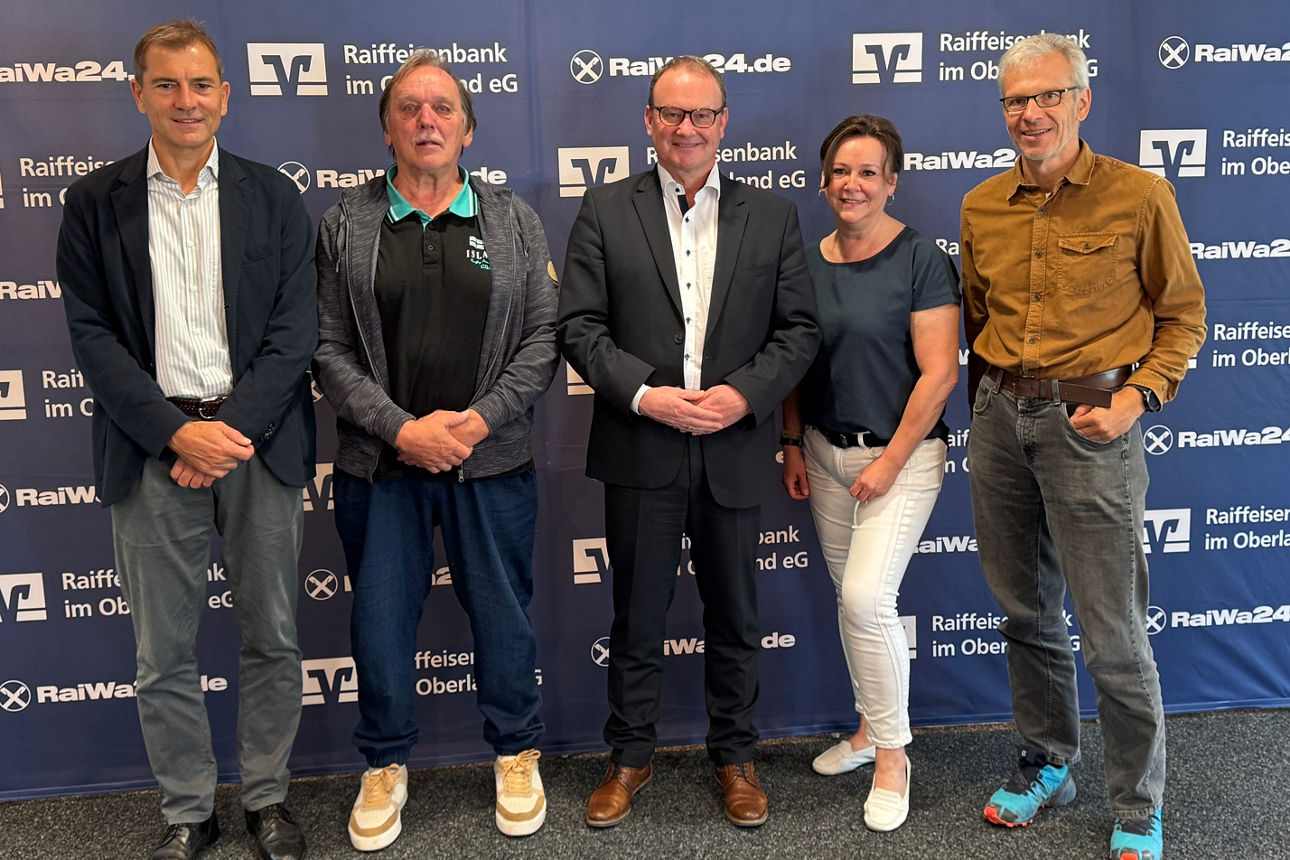 Fünf Personen (vier Männer, eine Frau) lächeln vor einer blauen Wand mit Raiffeisenbank-Logos.