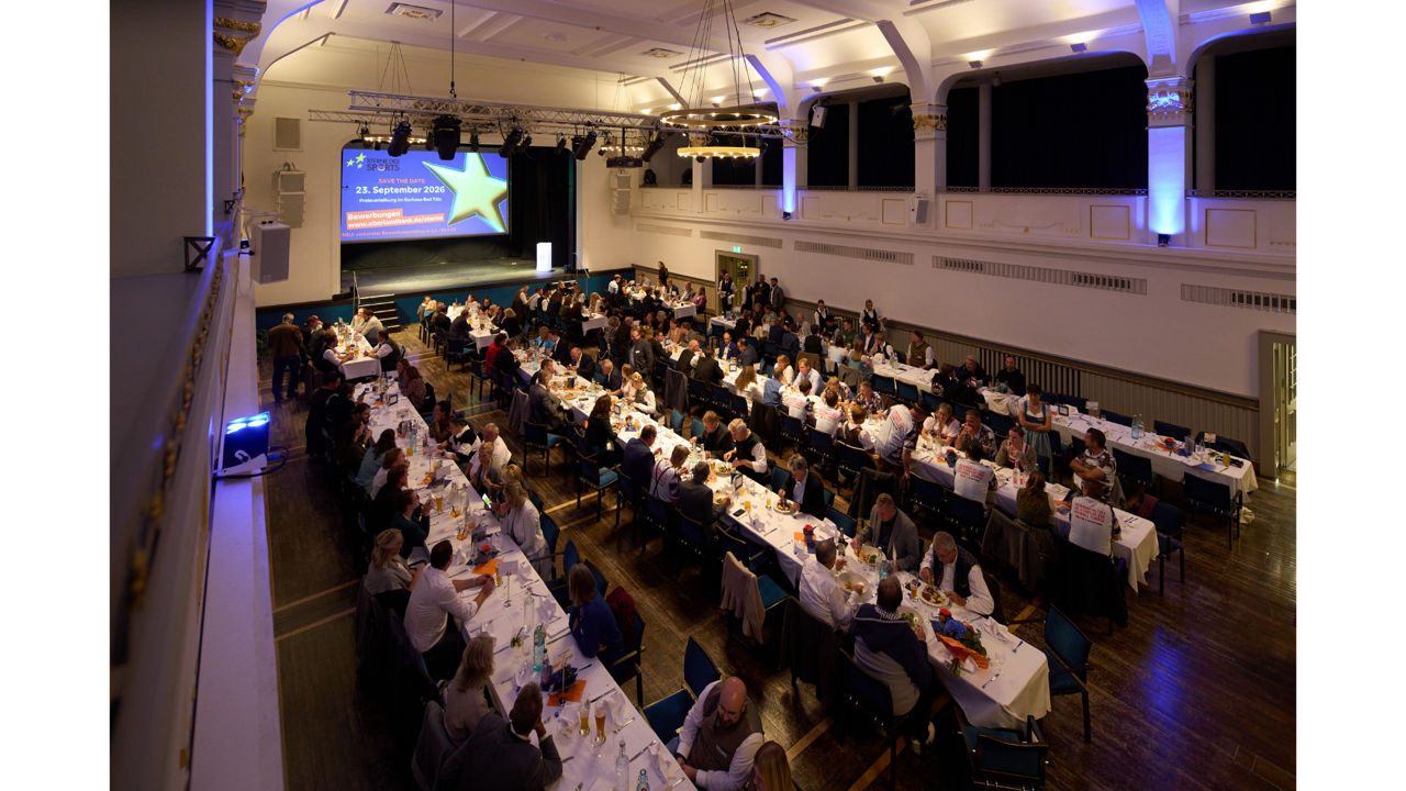 In einem festlichen Saal sitzen alle Gäste an langen in weiß eingedeckten Tafeln.