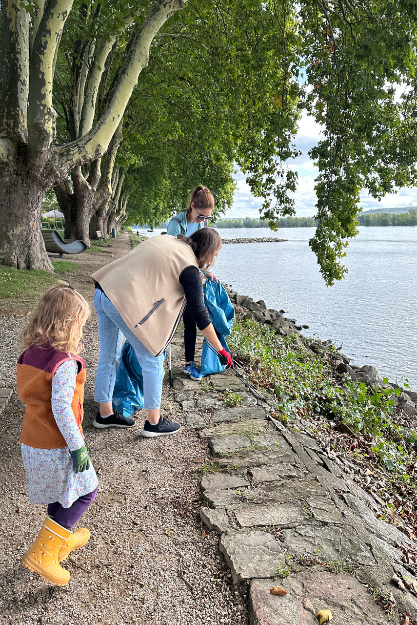 Drei Personen stehen am Ufer eines Gewässers. Die Personen sind dabei Müll einzusammeln. Im Hintergrund sind Bäume und Wasser sichtbar.