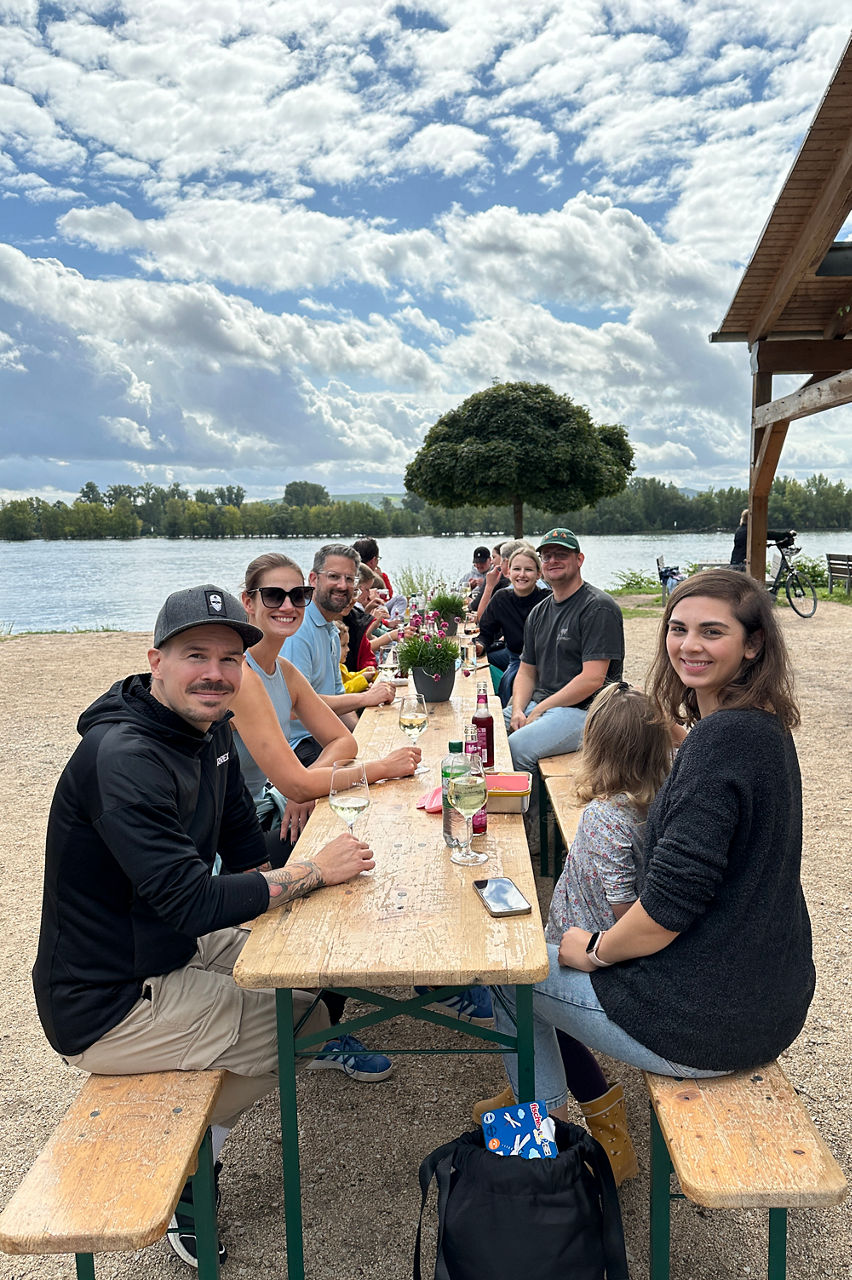 Eine Gruppe von mehreren Personen sitzt an einem langen Holztisch am Ufer eines Gewässers. Im Hintergrund sind Bäume und ein locker bewölkter blauer Himmel sichtbar.