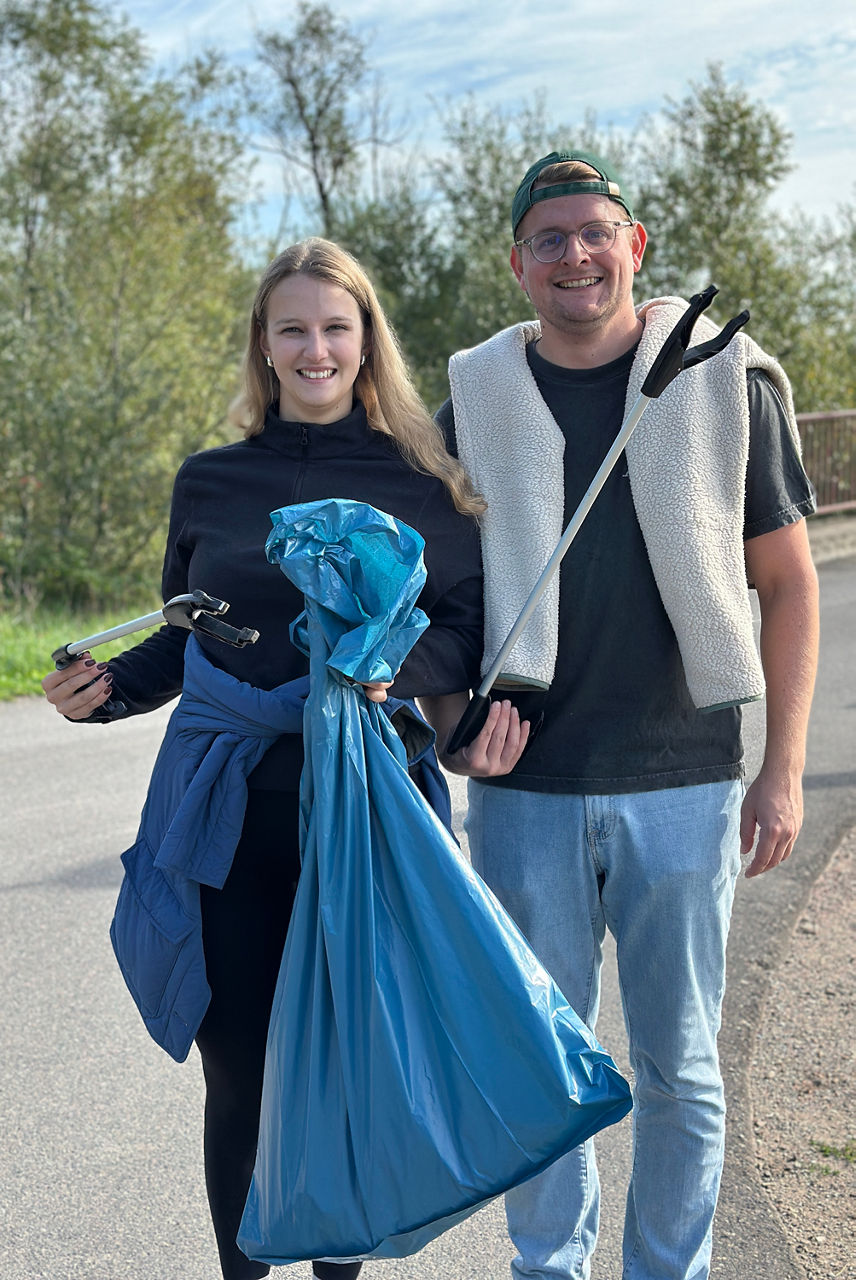 Zwei Personen stehen auf einem Weg. Eine Person hält einen blauen Müllsack und eine Greifzange, die andere eine Greifzange. Beide tragen legere Kleidung.