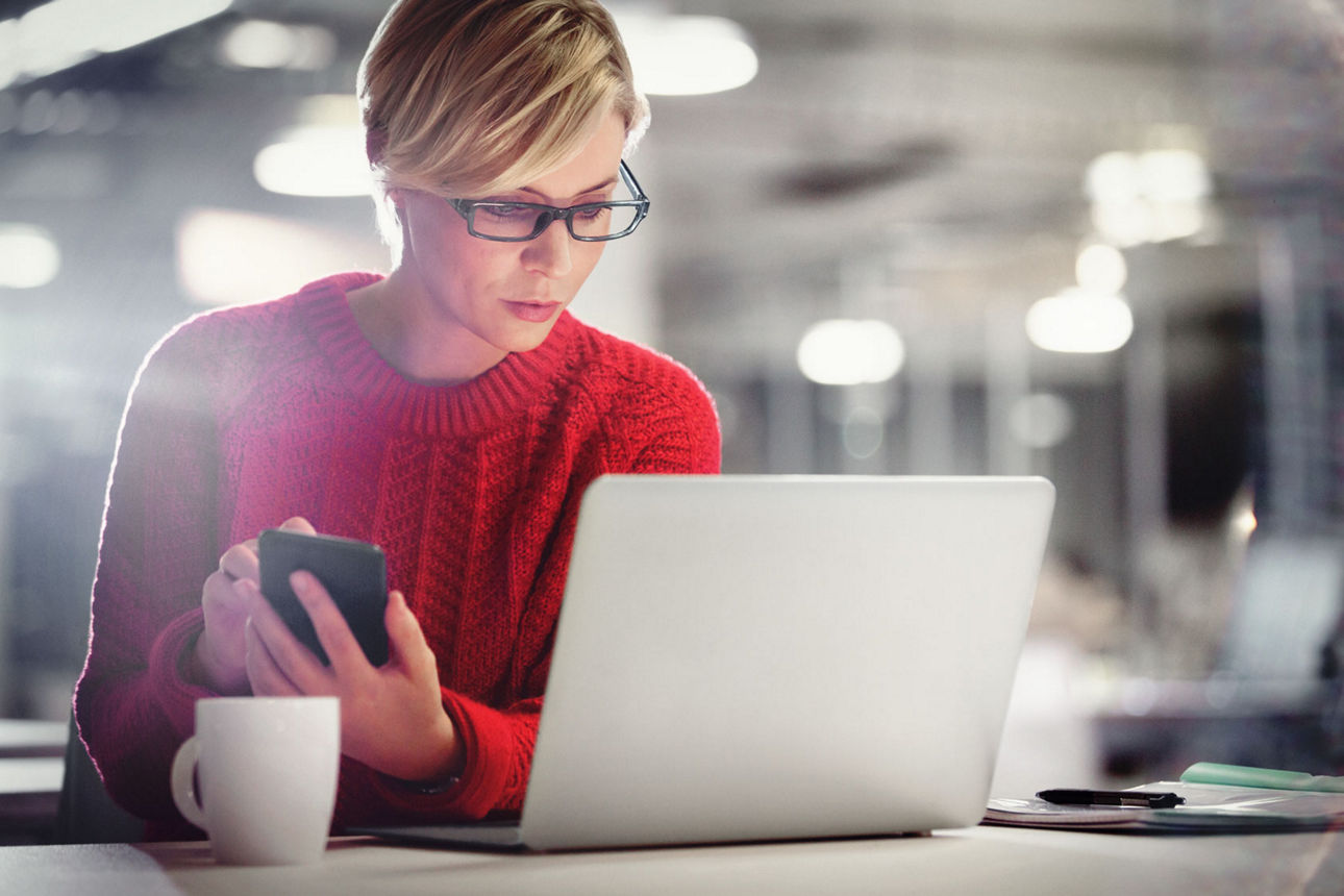 Frau im roten Pulli sitzt mit einem Smartphone vor einem Laptop