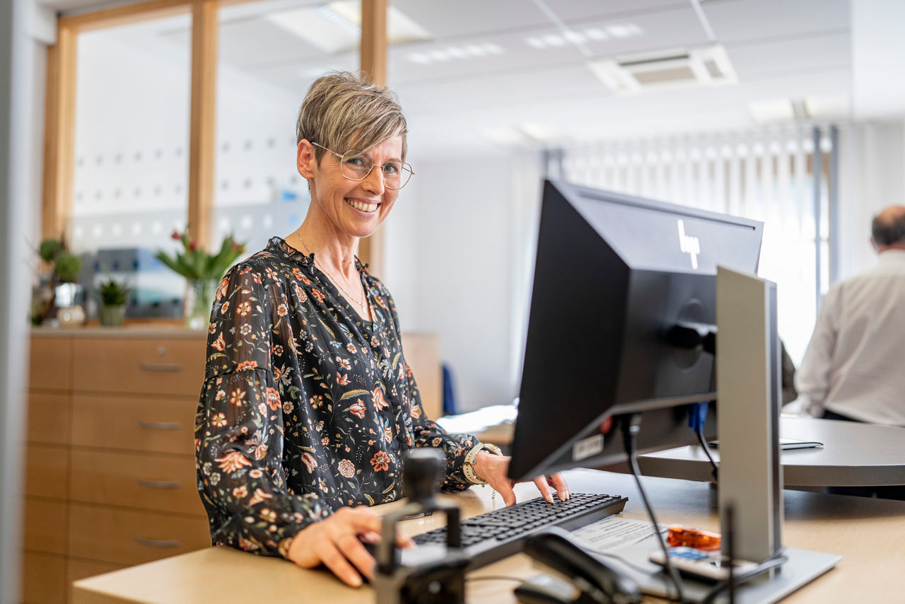 Eine lächelnde Frau steht vor einem PC an ihrem Arbeitsplatz