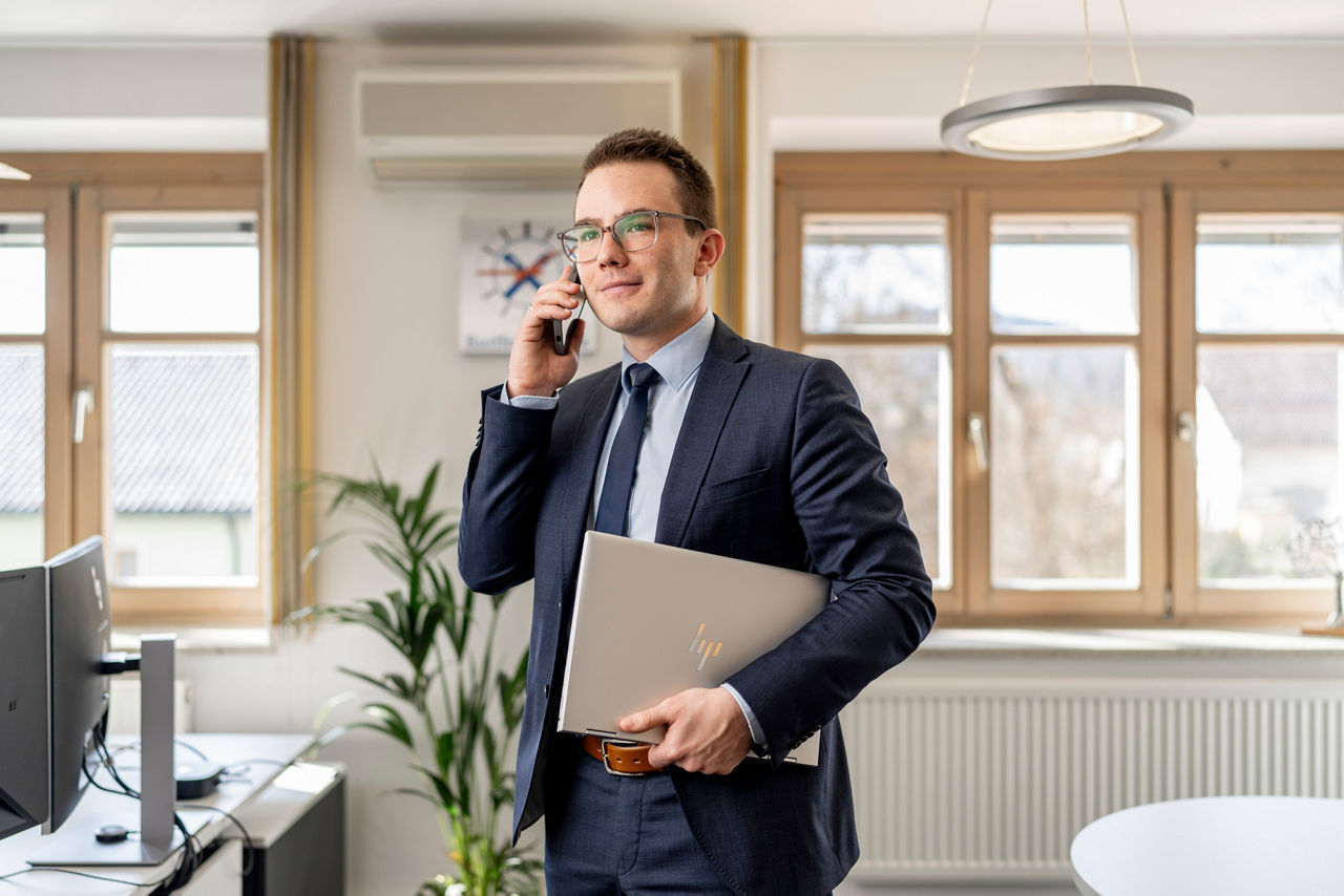 ein junger Herr im Anzug steht mit Laptop im Arm und Handy am Ohr in einem hellen Büro