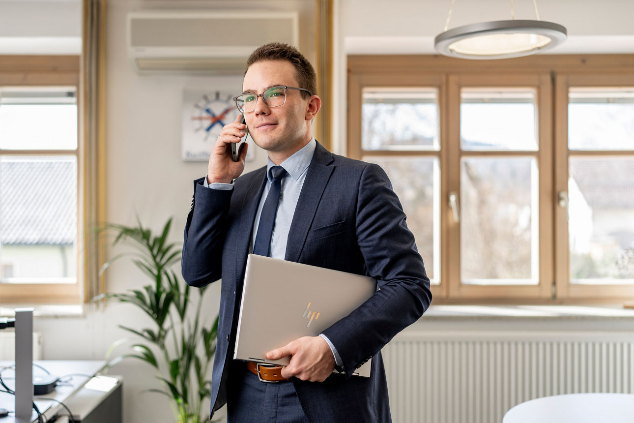 Mann mit Anzug mit Handy am Ohr und Laptop in der Hand in einem Büro