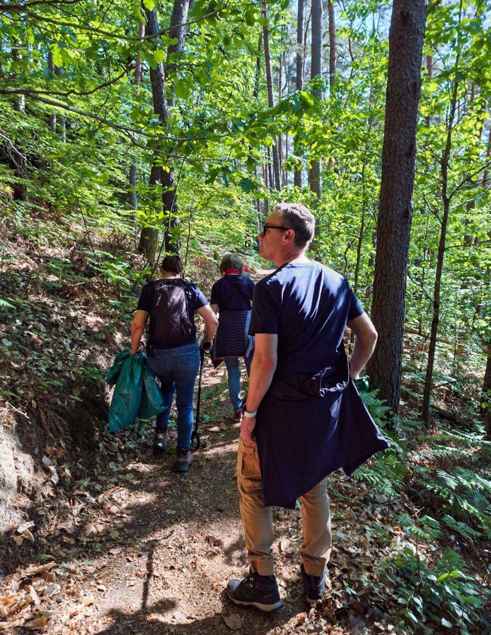 Drei Personen gehen auf einem schmalen Waldweg bergauf und tragen Säcke bei einer gemeinsamen Aktion