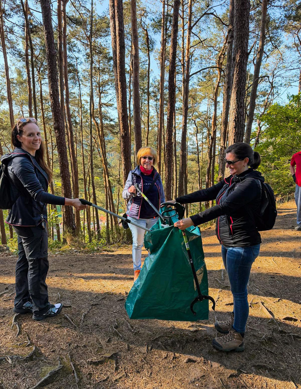 Drei Personen halten gemeinsam einen großen grünen Müllsack und nutzen Greifzangen auf einem Waldweg.