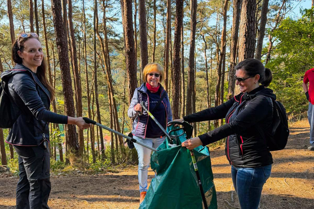 Drei Personen halten gemeinsam einen großen grünen Müllsack und nutzen Greifzangen auf einem Waldweg.
