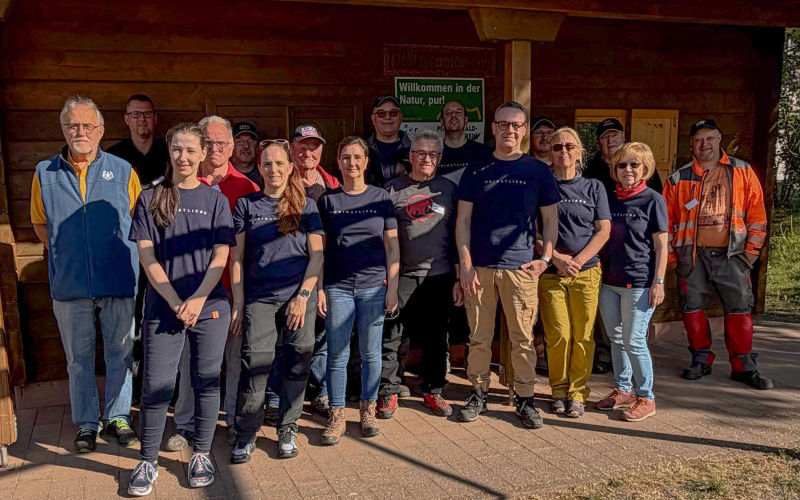 Gruppe von Personen posiert vor einem Holzgebäude; einige tragen passende T‑Shirts.