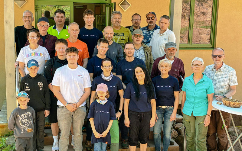 Gruppe von Personen posiert vor einem Gebäudeeingang; einige tragen gleiche T‑Shirts, ein Tisch steht rechts