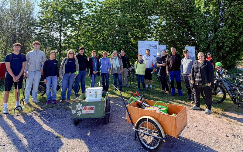 Gruppe steht im Freien vor Infotafeln, davor Handwagen und Fahrräder mit Arbeitsgeräten.