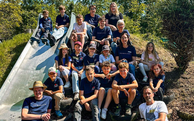 Gruppe aus Kindern und Erwachsenen sitzt und steht an einer Metallrutsche in einem parkähnlichen Gelände.