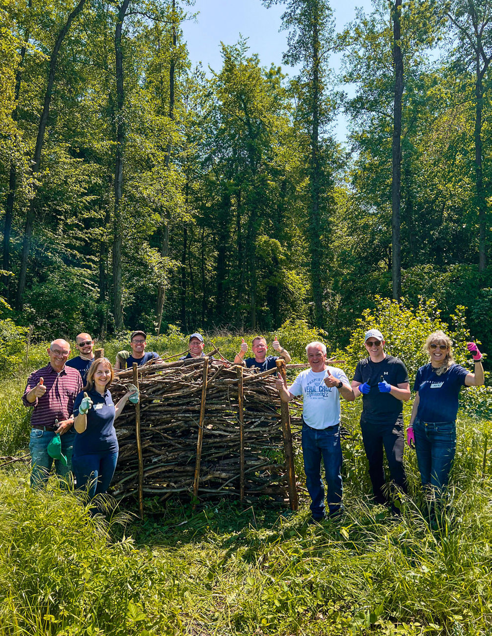 Mehrere Personen stehen im Wald vor einem Zaun aus Ästen. Sie zeigen Daumen hoch. Rundherum ist viel Grün.